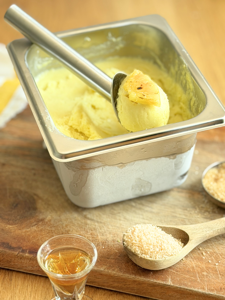 sorbet pina colada dessert glace deliciouslyhome article2 Sorbet Piña Colada crémeux garni d'un morceau d'ananas rôti, présenté dans un bac inox avec noix de coco râpée et rhum en accompagnement. Découvrez la recette complète en lisant notre article sur Deliciously Home.
