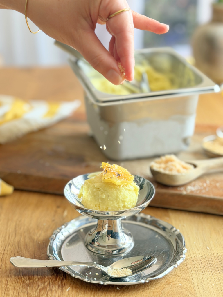 sorbet pina colada dessert glace deliciouslyhome article3 Main saupoudrant de la noix de coco râpée sur une coupe de sorbet Piña Colada garnie d'un morceau d'ananas rôti. Découvrez la recette complète en lisant notre article sur Deliciously Home.