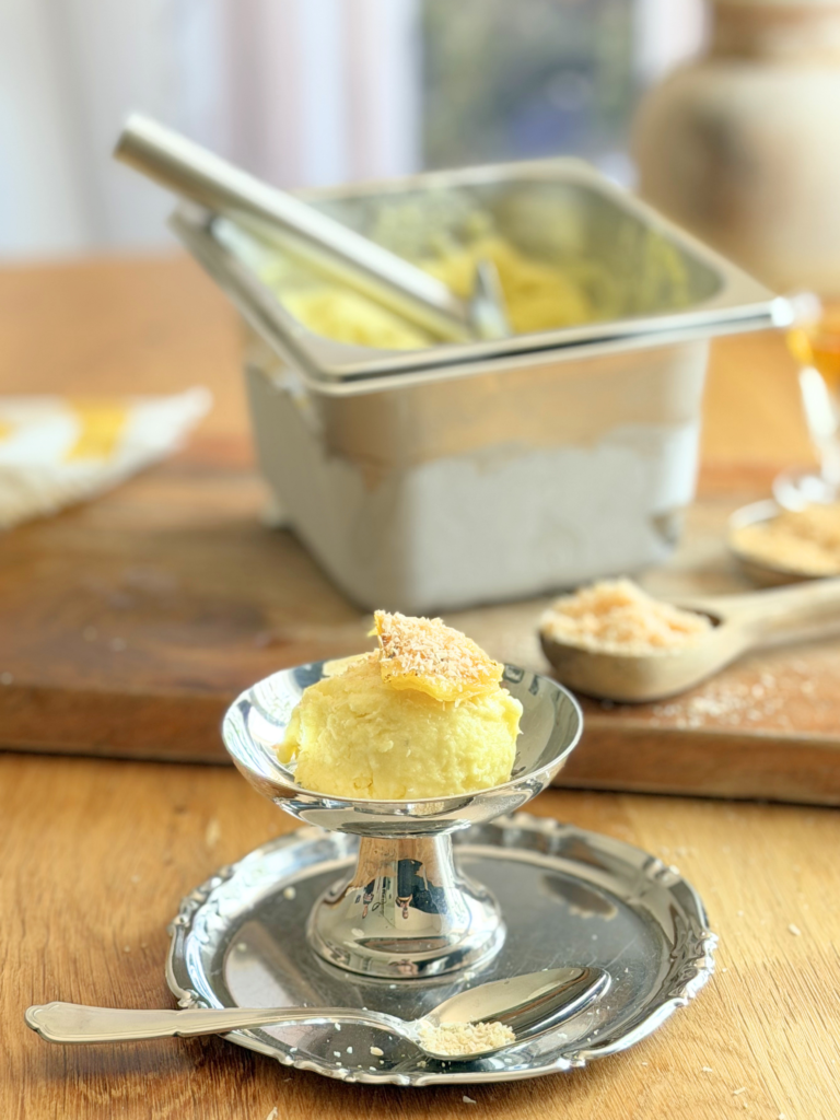 sorbet pina colada dessert glace deliciouslyhome article4 Coupe de sorbet Piña Colada maison décorée d'un morceau d'ananas rôti et de noix de coco râpée, servie sur un plateau en métal. Découvrez la recette complète en lisant notre article sur Deliciously Home.