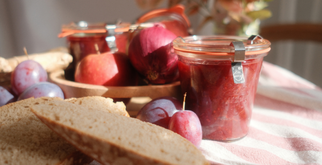 Pots de chutney de prunes maison entourés de prunes et d’un oignon rouge, dans une lumière automnale, recette Deliciously Home.