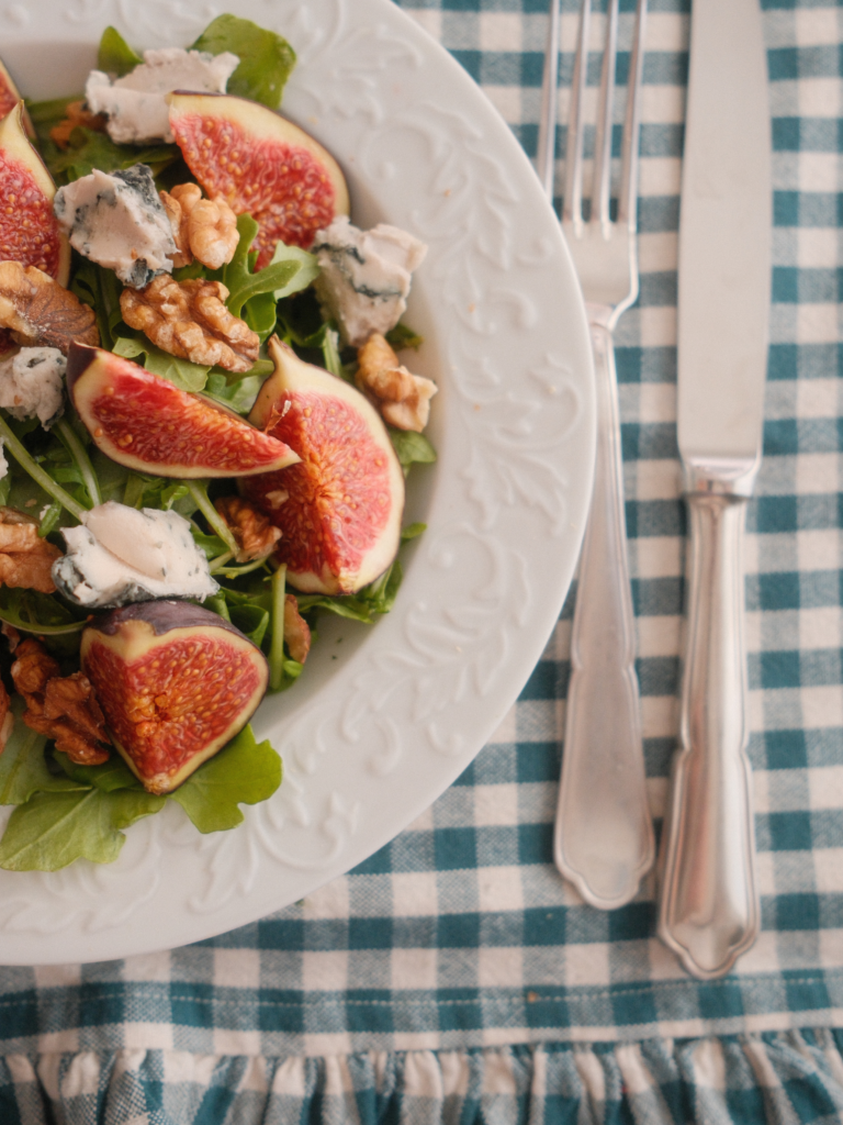 Salade de figues violettes et fromage bleu avec cresson et noix, servie dans une assiette blanche sur une nappe à carreaux. Retrouvez la recette sur Deliciously Home.
