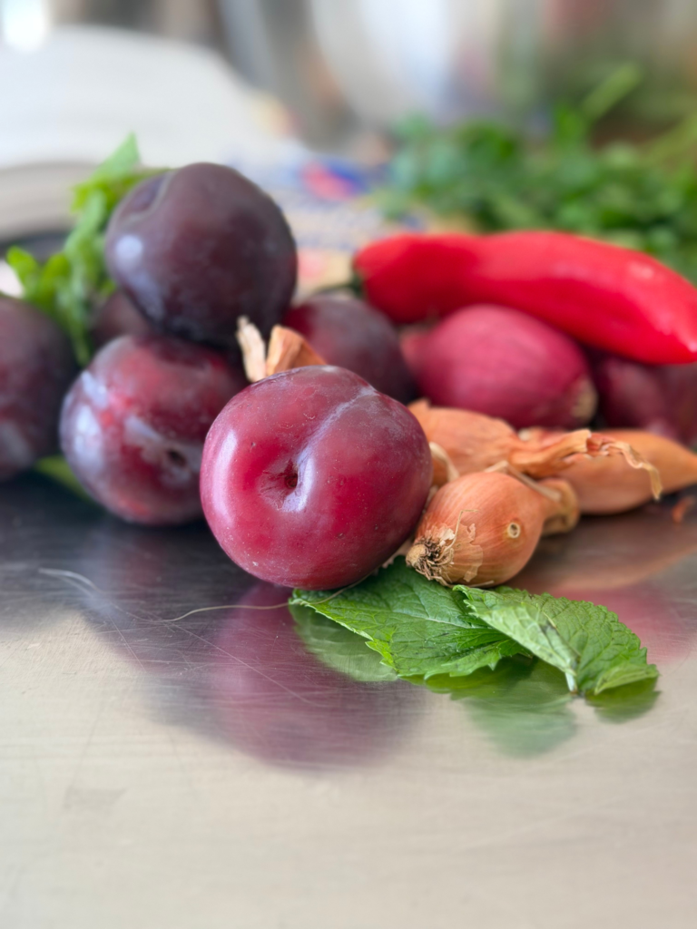 Prunes violettes de saison entourées d’échalotes, d’oignons rouges, de menthe et d’un piment rouge avant la préparation du taboulé. Retrouvez la recette sur Deliciously Home.